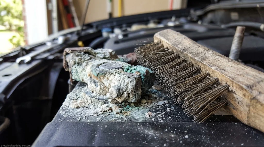 Close-up of white and blue-green corrosion on a car battery terminal next to a wire brush