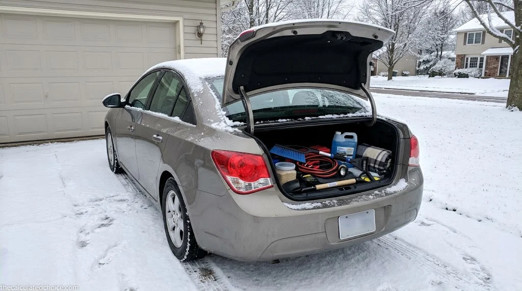 How to winterize your car: sedan in a snowy driveway with the trunk open, packed with jumper cables, washer fluid, a snow brush and a blanket